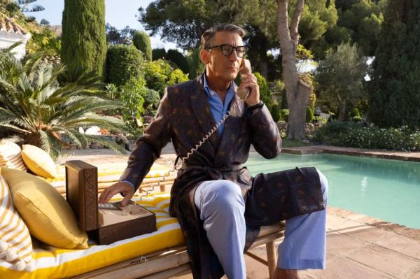 Jason Issacs sitting next to a pool holding a phone. 