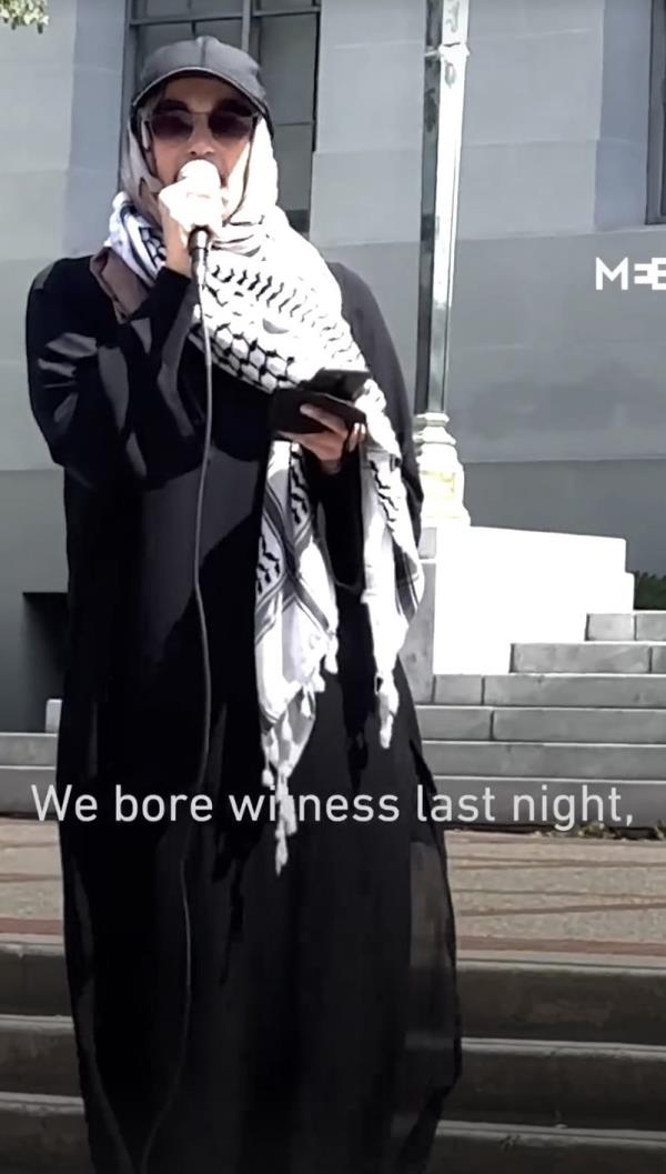Malak Afaneh in ba<em></em>seball cap and a hijab made out of a kefiyeh, and a long black robe, holding a microphone.
