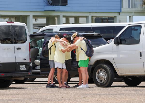 Teens in yellow shirts stand in a circle with their arms around each other and their heads bowed down.