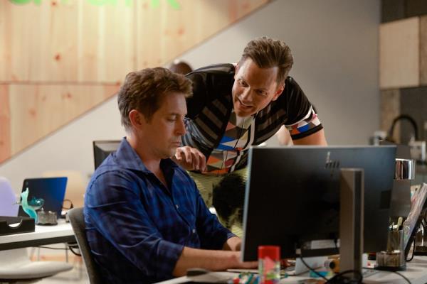 Beck Bennett sitting at a computer while Joel McHale leans over him. 