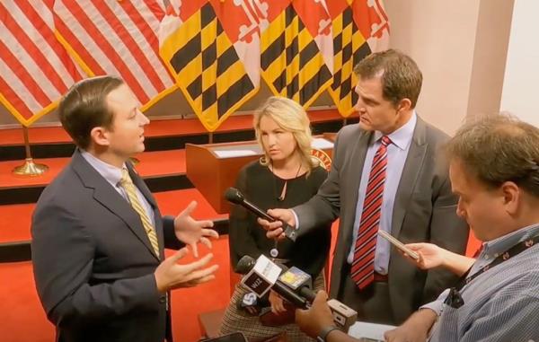 Man in suit answering reporters questions in front of Maryland flags