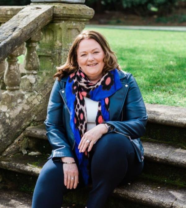 Alison Rios McCrone sitting outside on some stone steps, with a stone balustrade on her left and grass behind her.
