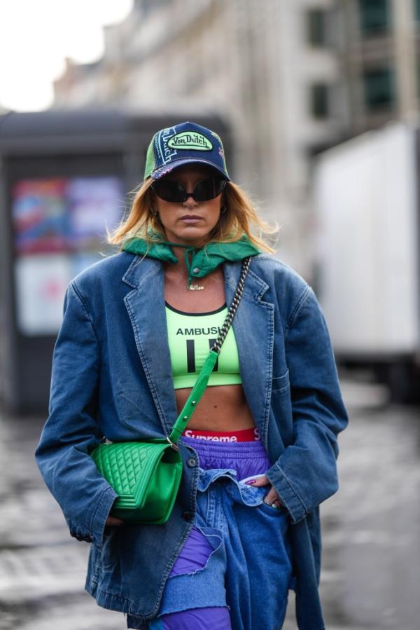 A fashio<em></em>nably dressed guest at Paris Fashion Week, wearing a blue denim and green mesh cap, black sunglasses, a green hoodie, gold necklaces, and a blue denim jacket, holding a green Chanel crossbody bag.