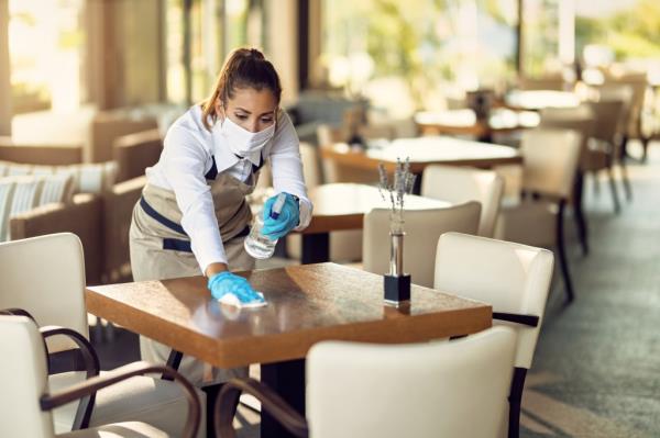 Young waitress wearing a face mask and gloves while disinfecting tables in a café during the COVID-19 pandemic.