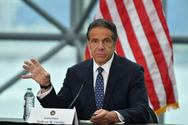 Former New York Gov. Andrew Cuomo gestures while seated at a table with a placard bearing his name in front of him and an American flag in the background. 
