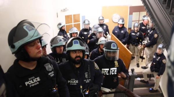 Police in riot gear marked NYPD on stairs inside a building.