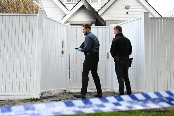 Two police detectives stand outside a tall white gate behind police tape
