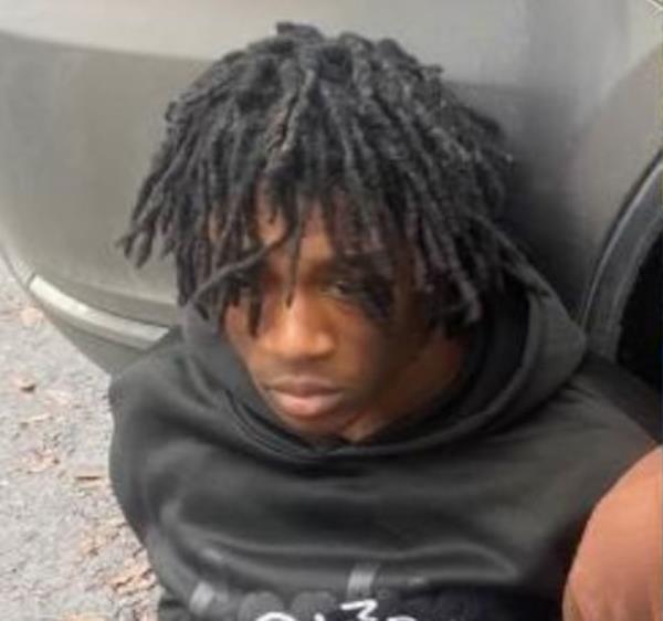 A young man with a black hoodie and short dreads sitting on the ground leaning against the rear bumper of a car.
