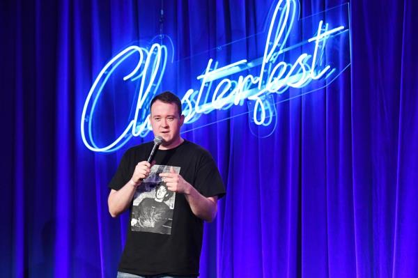 Shane Gillis speaking into a microphone in front of a neon light that says Clusterfest in cursive