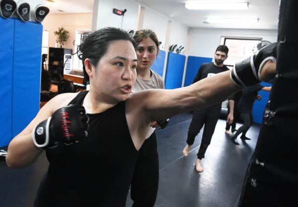 Woman in black shirt and boxing gloves punches a bag while others look on.