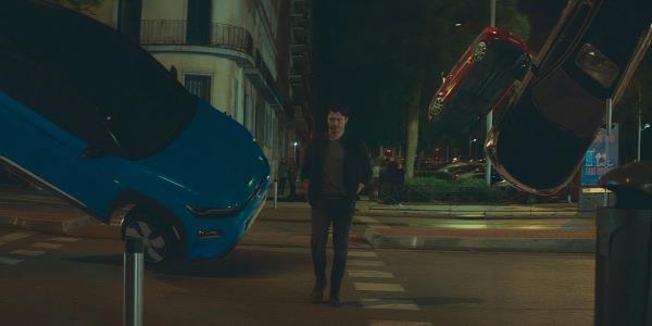 Henry Golding walks through a street with flipping cars in Daniela Forever