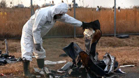 EPA A member of Korea Crime Scene Investigation in a white hazmat suit inspects the wreckage of the Jeju Air aircraft at Muan Internatio<em></em>nal Airport. They are holding up what appears to be the remains of a plane seat