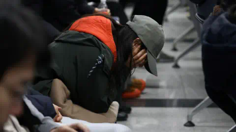 Getty Images A woman sits on a row of seats at an airport with her head in her hands. She is wearing a ba<em></em>seball cap and you can't see her face